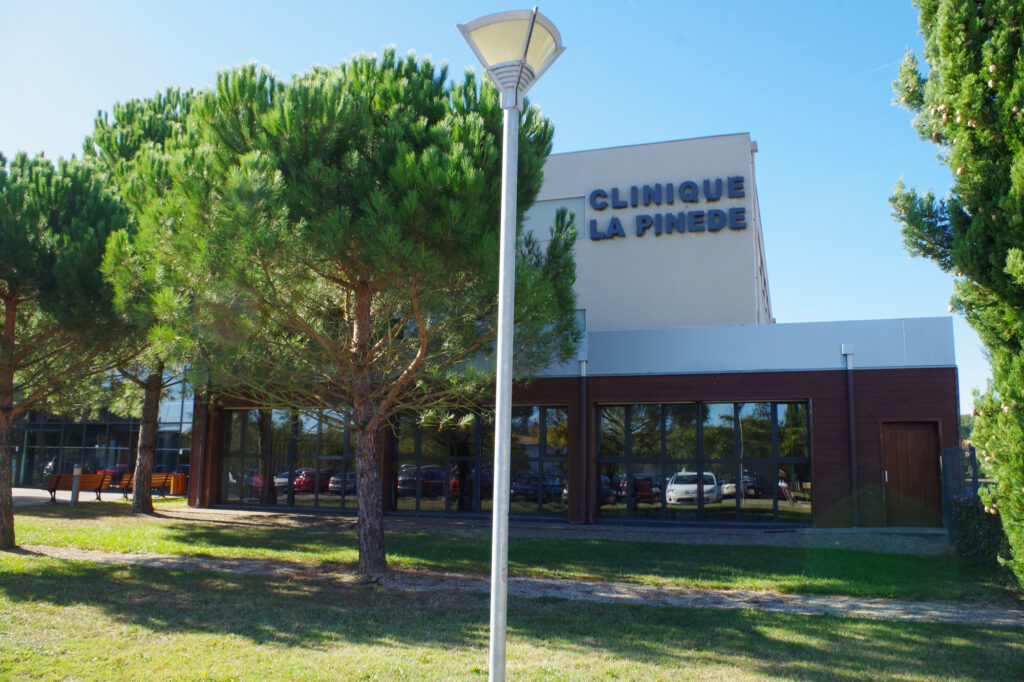 Vue sur l'enseigne de la Clinique La Pinède depuis le parking. Le bâtiment principal est partiellement visible avec des arbres, des pins, à l'avant.