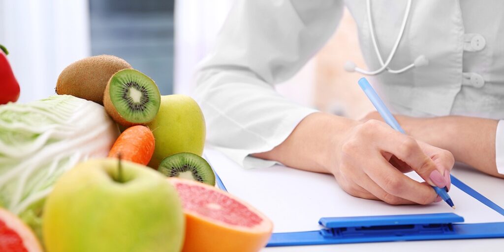 Gros plan sur l'avant-bras et la main d'une diététicienne en train de rédiger une ordonnance. À côté de sa main, des fruits et légumes frais, tranchés en deux ou entier.